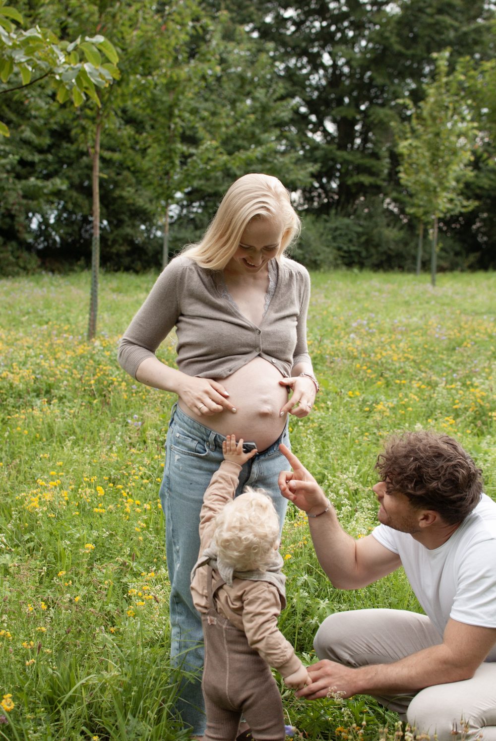 Familie auf der Blumenwiese: Mama zeigt auf den Bauch, das Kleinkind greift danach, Papa hockt daneben