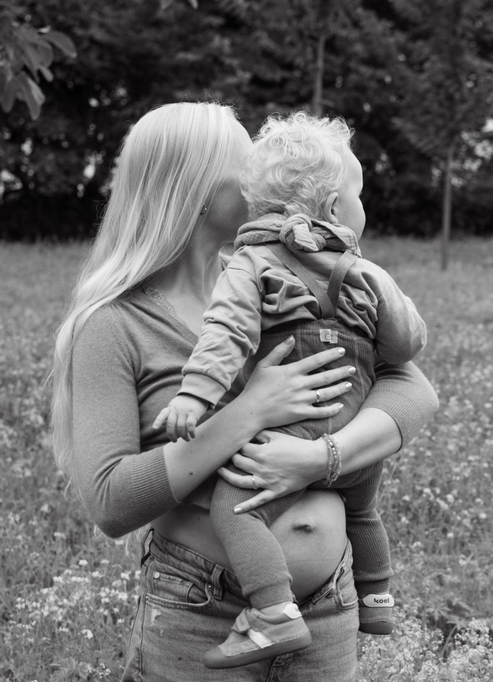 Schwarz-weiß: Die Mama trägt ihr Kleinkind auf dem Arm und küsst es, der Babybauch ist zu sehen