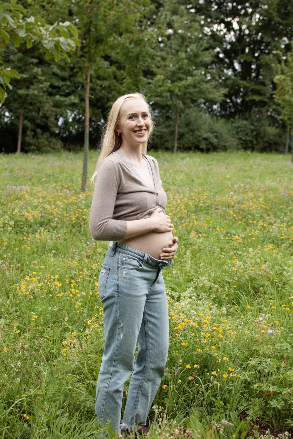 Lachende Schwangere in Jeans und Strickjacke auf einer Blumenwiese, die Hände am Babybauch