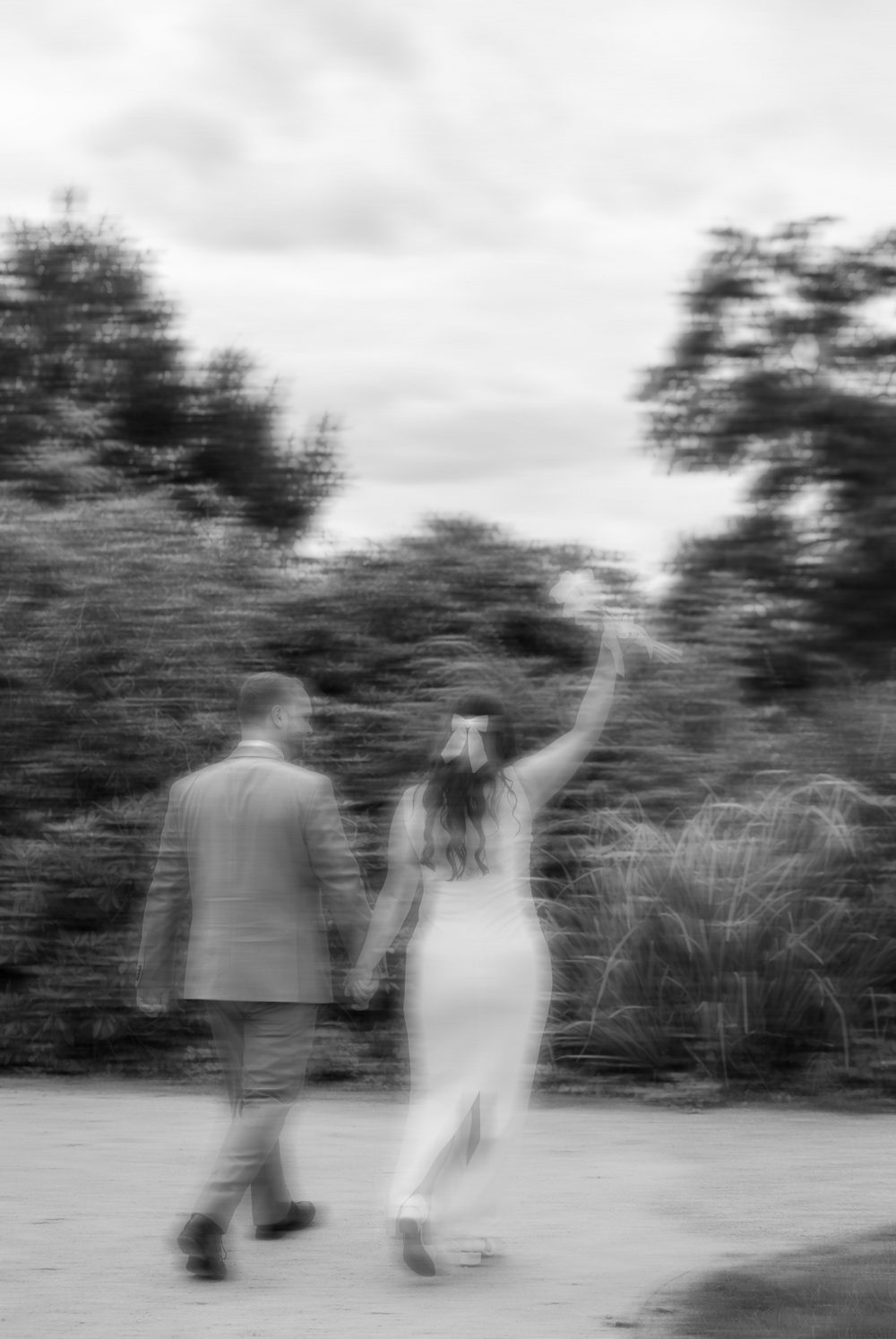 Schwarz-weiß: Das Brautpaar geht Hand in Hand einen Parkweg entlang, sie reckt den Strauß in die Luft